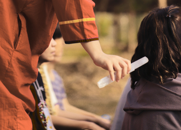 A healer gently working with a selenite wand on a seated person outdoors — one-on-one session grounded in embodied attention and care